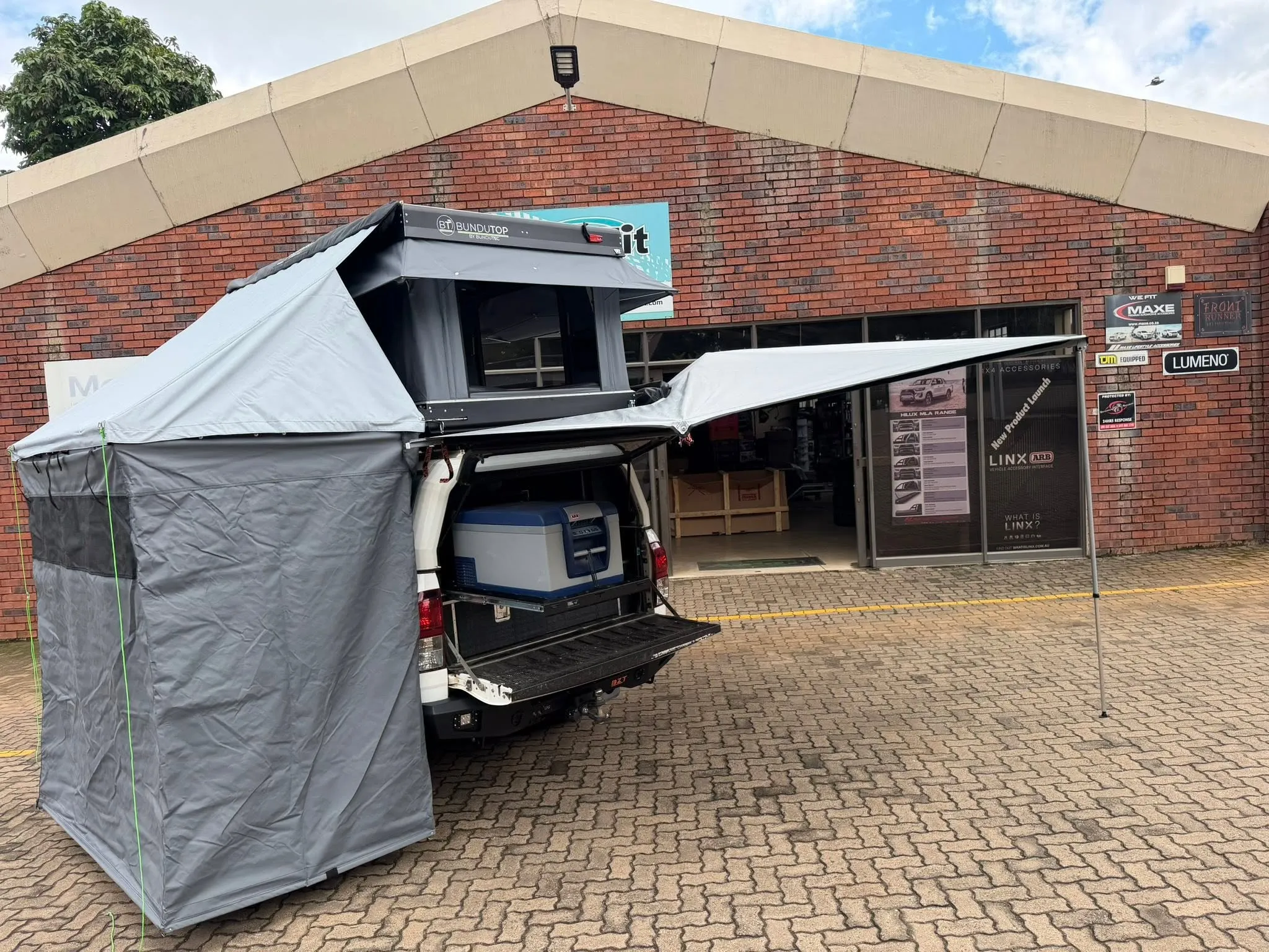 White Toyota Hilux double cab fitted with a BunduTop electric popup rooftop tent, side awning, and an ARB camping fridge on a slide-out