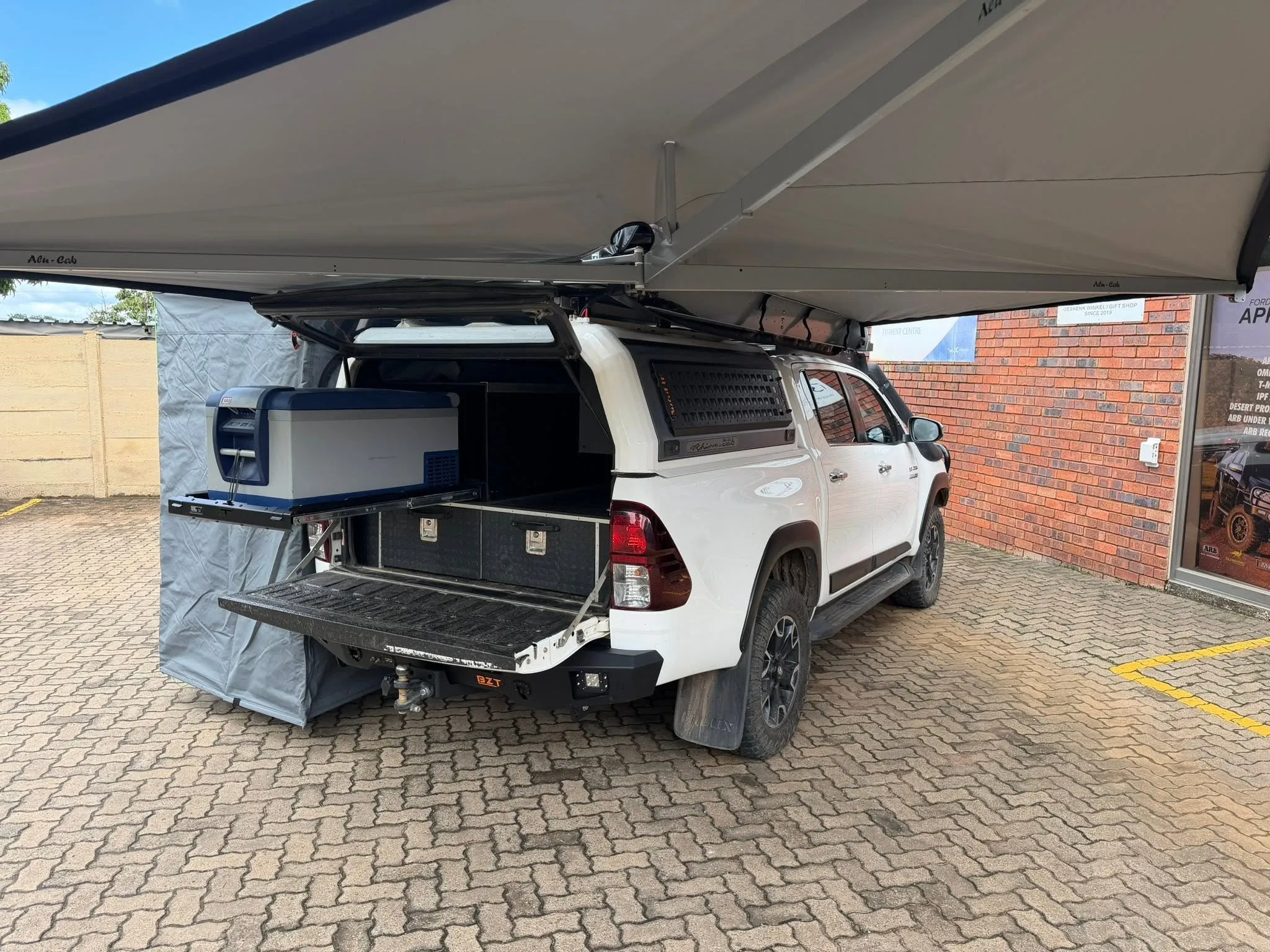 White Toyota Hilux double cab fitted with an Alu-Cab 270-degree awning, Rhino Cab canopy, and an ARB camping fridge on a slide-out drawer system