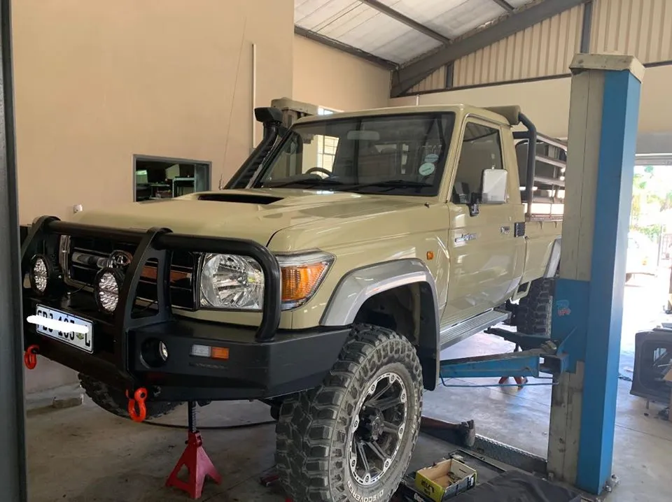 Sand-colored Toyota Land Cruiser 79 single-cab on a workshop lift showing heavy-duty steel replacement front bumper, round LED spotlights, and snorkel