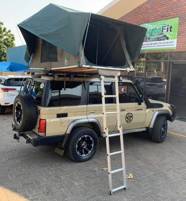 Side view of a beige Toyota Land Cruiser 76 Series station wagon with an open soft-shell rooftop tent and extension ladder parked in front of the AutoFit building