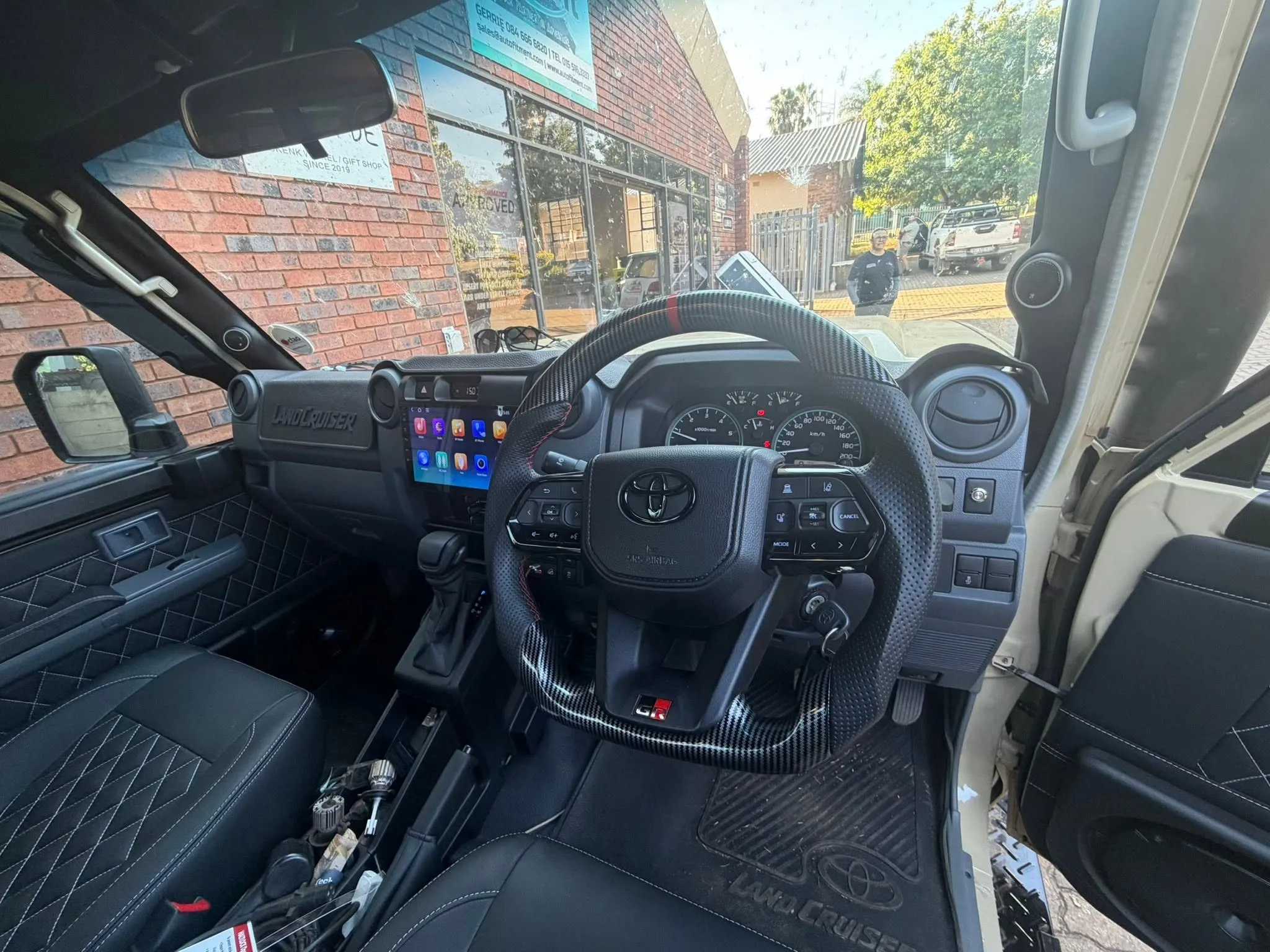 Interior dashboard view of a Toyota Land Cruiser 70 Series showing custom carbon-fiber steering wheel, diamond-stitched leather seats, and touchscreen infotainment unit