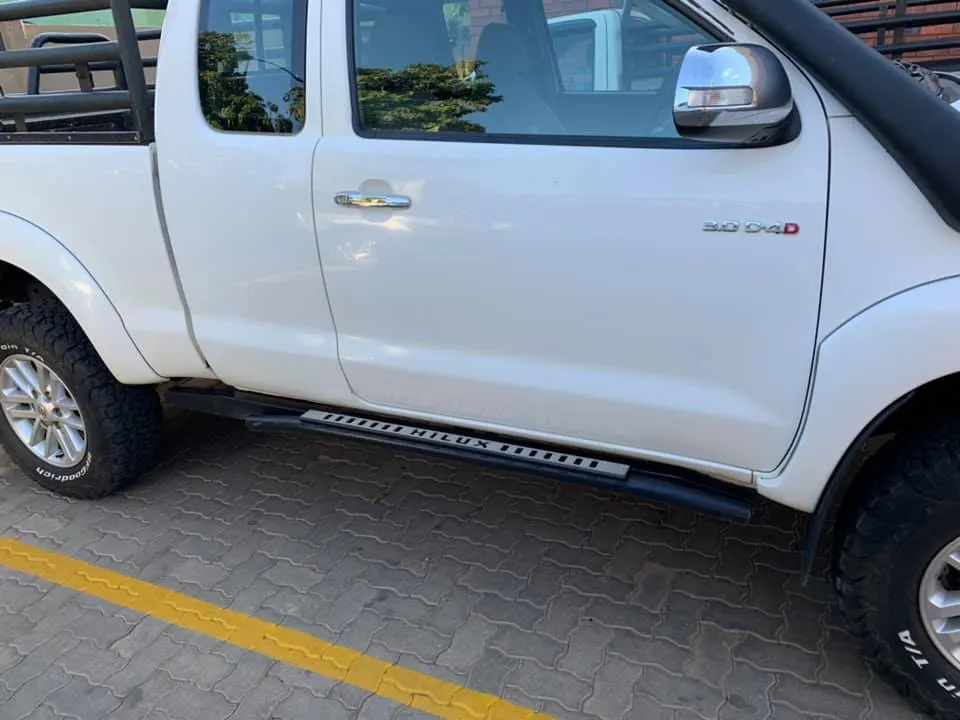 Side view of a white Toyota Hilux 3.0 D-4D showing custom matte black side steps with Hilux lettering, A-pillar snorkel, and BFGoodrich All-Terrain tyres
