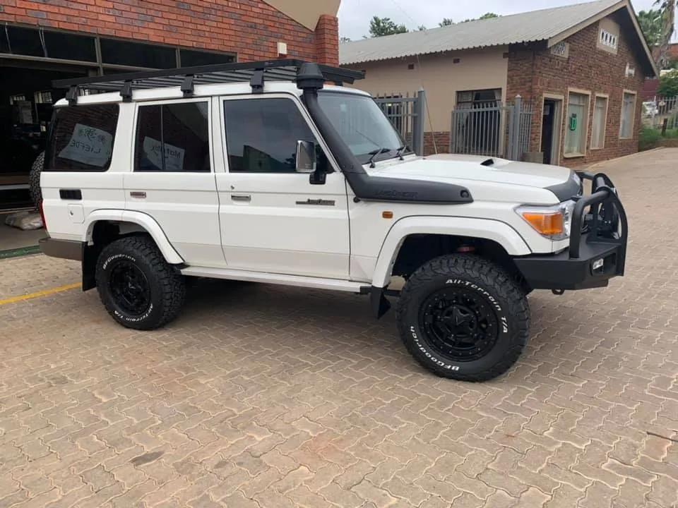 White Toyota Land Cruiser 76 station wagon side view showing a low-profile roof rack, steel bumper, and off-road wheels